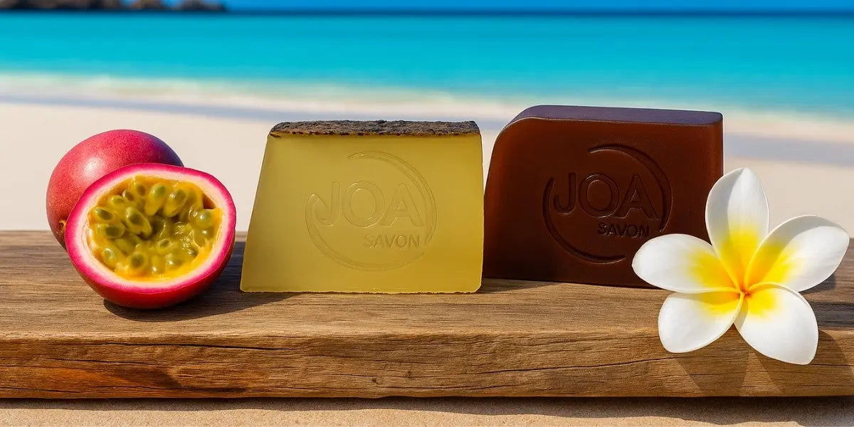 Savons Joa du mois d’août : savon Monoï et savon Fruit de la Passion posés sur une plage, avec fleur de tiaré et fruits exotiques en décor naturel