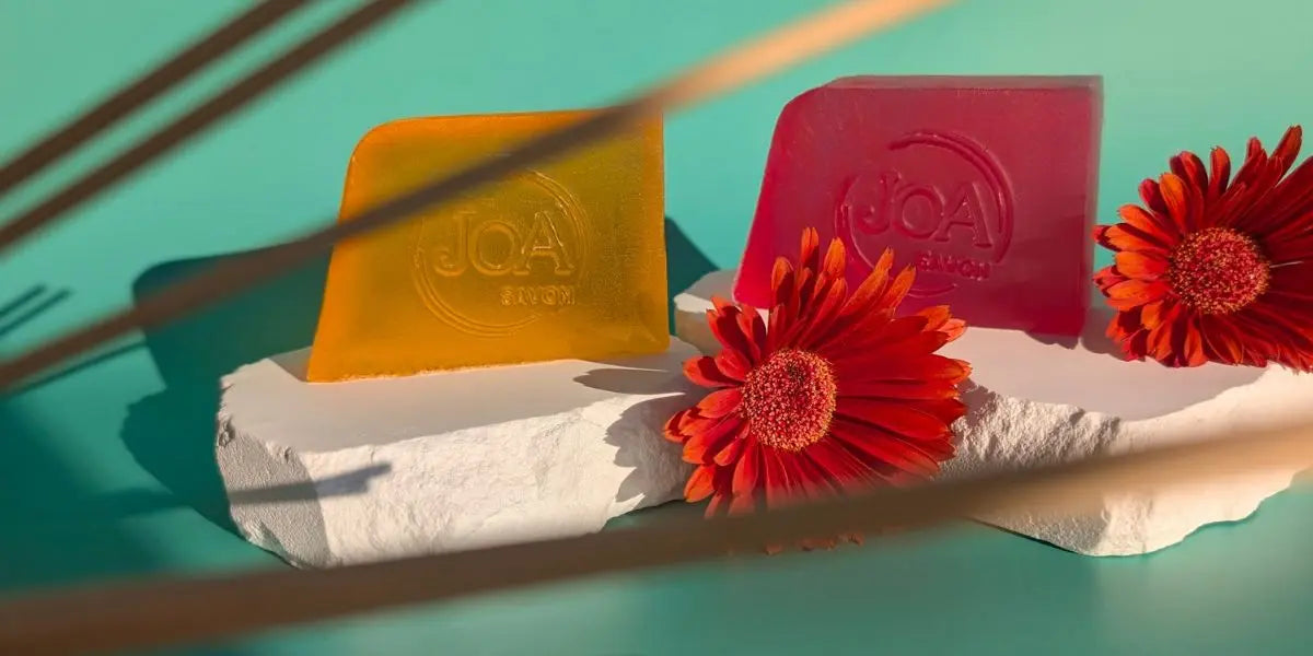 Deux savons Joa translucides orange et rose posés sur un socle blanc, avec deux fleurs rouges sur fond turquoise ensoleillé