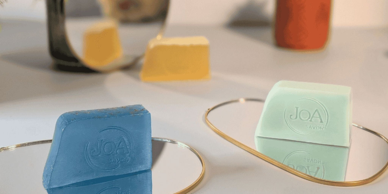 Savons Les Incontournables Joa Savon posés sur miroirs dorés, ambiance douce et lumineuse avec plantes et reflets, savons bleus, verts et jaunes