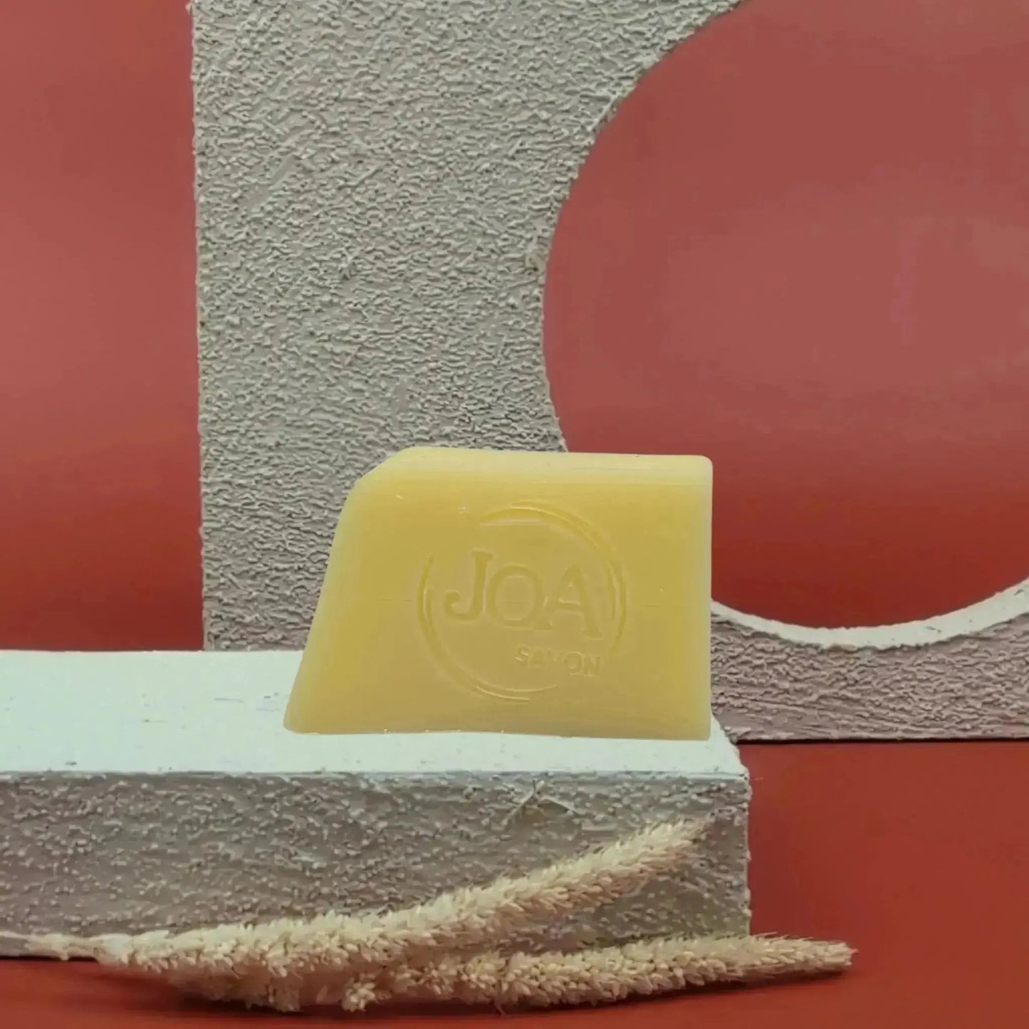 Savon solide Abricot Joa jaune pâle, posé sur socle blanc texturé avec fond rouge et décor géométrique