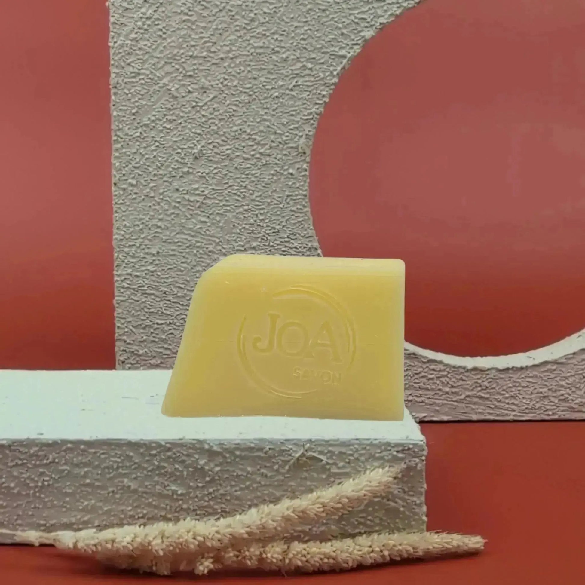 Savon solide Abricot Joa jaune pâle, posé sur socle blanc texturé avec fond rouge et décor géométrique