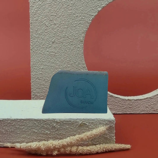 Savon solide Algues Joa bleu profond, posé sur socle blanc texturé avec fond rouge et structure géométrique