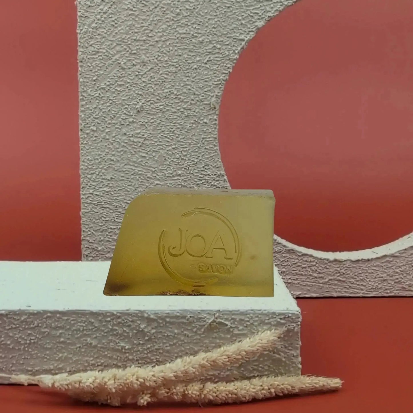 Savon solide Aloé-vera Joa translucide vert doré, posé sur socle blanc texturé avec fond rouge et formes géométriques