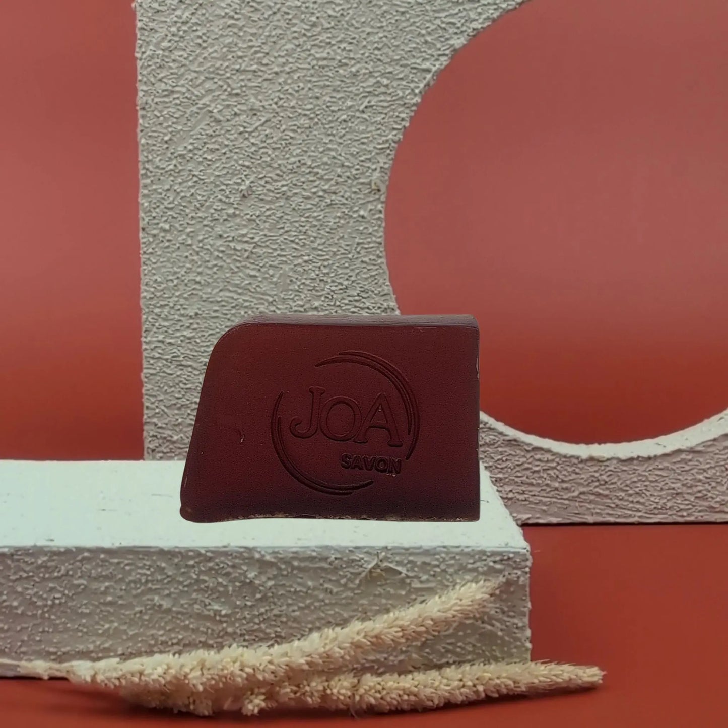 Savon solide Amarante Joa bordeaux profond, posé sur socle blanc texturé avec fond rouge et éléments géométriques