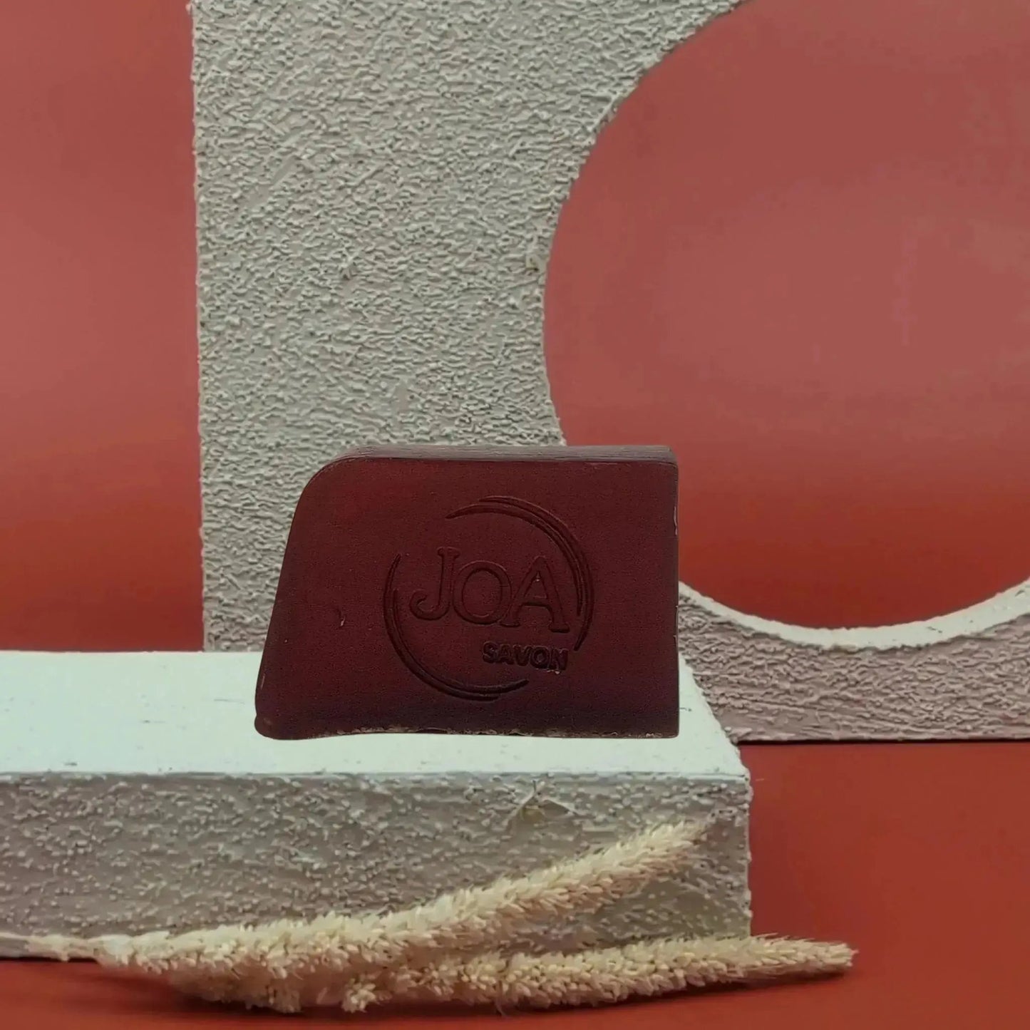 Savon solide Amarante Joa bordeaux profond, posé sur socle blanc texturé avec fond rouge et éléments géométriques