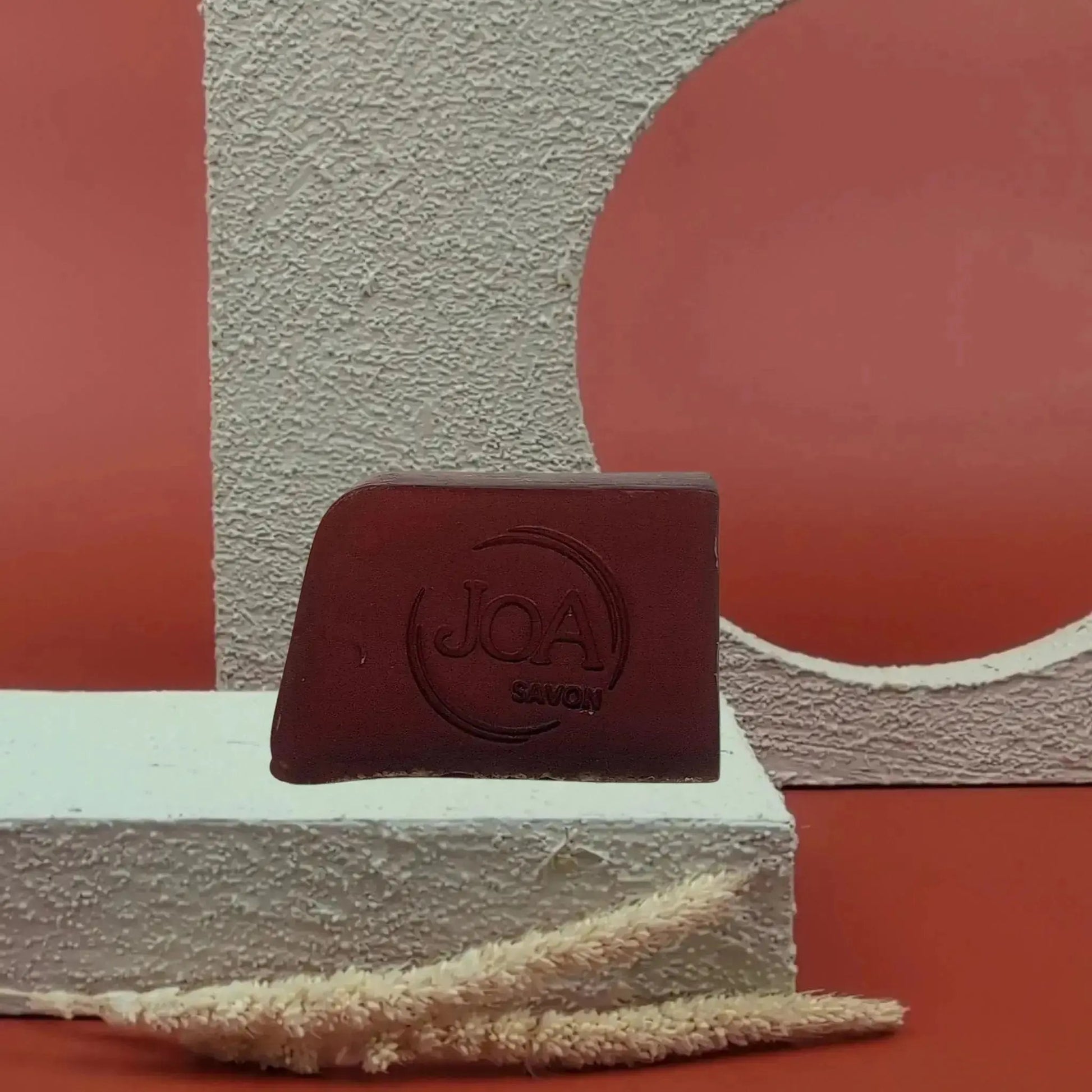 Savon solide Amarante Joa bordeaux profond, posé sur socle blanc texturé avec fond rouge et éléments géométriques