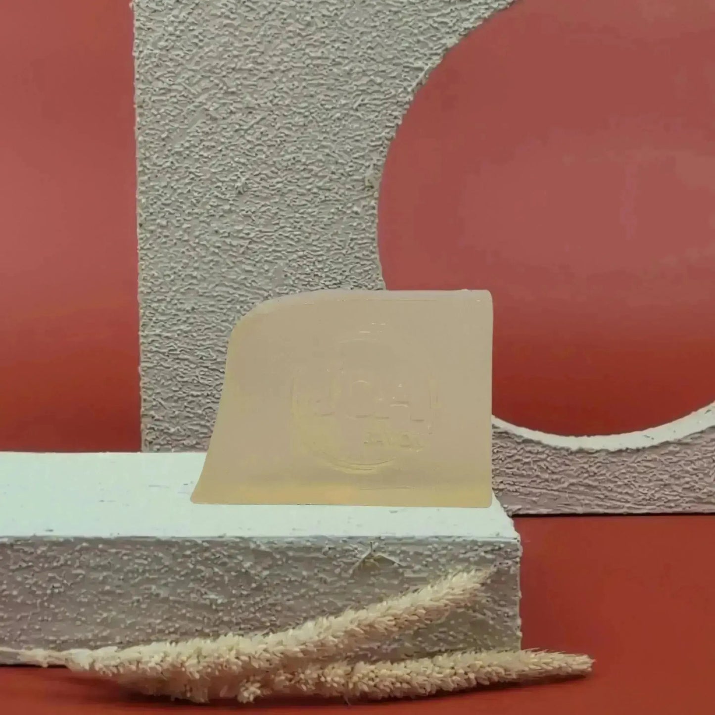 Savon solide Argan Joa beige doré, posé sur socle blanc texturé avec fond rouge et structure géométrique