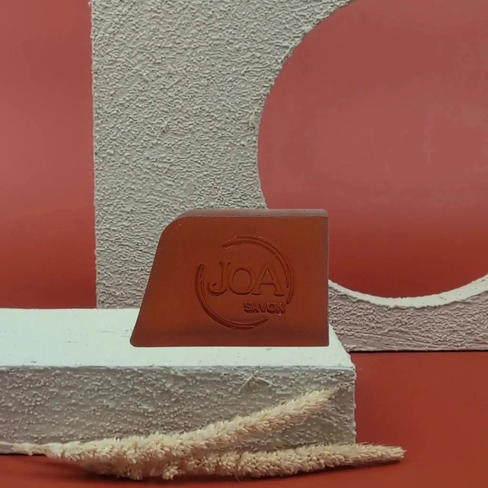 Savon solide Bella Vita Joa rouge terracotta, posé sur socle blanc texturé avec fond rouge et formes géométriques