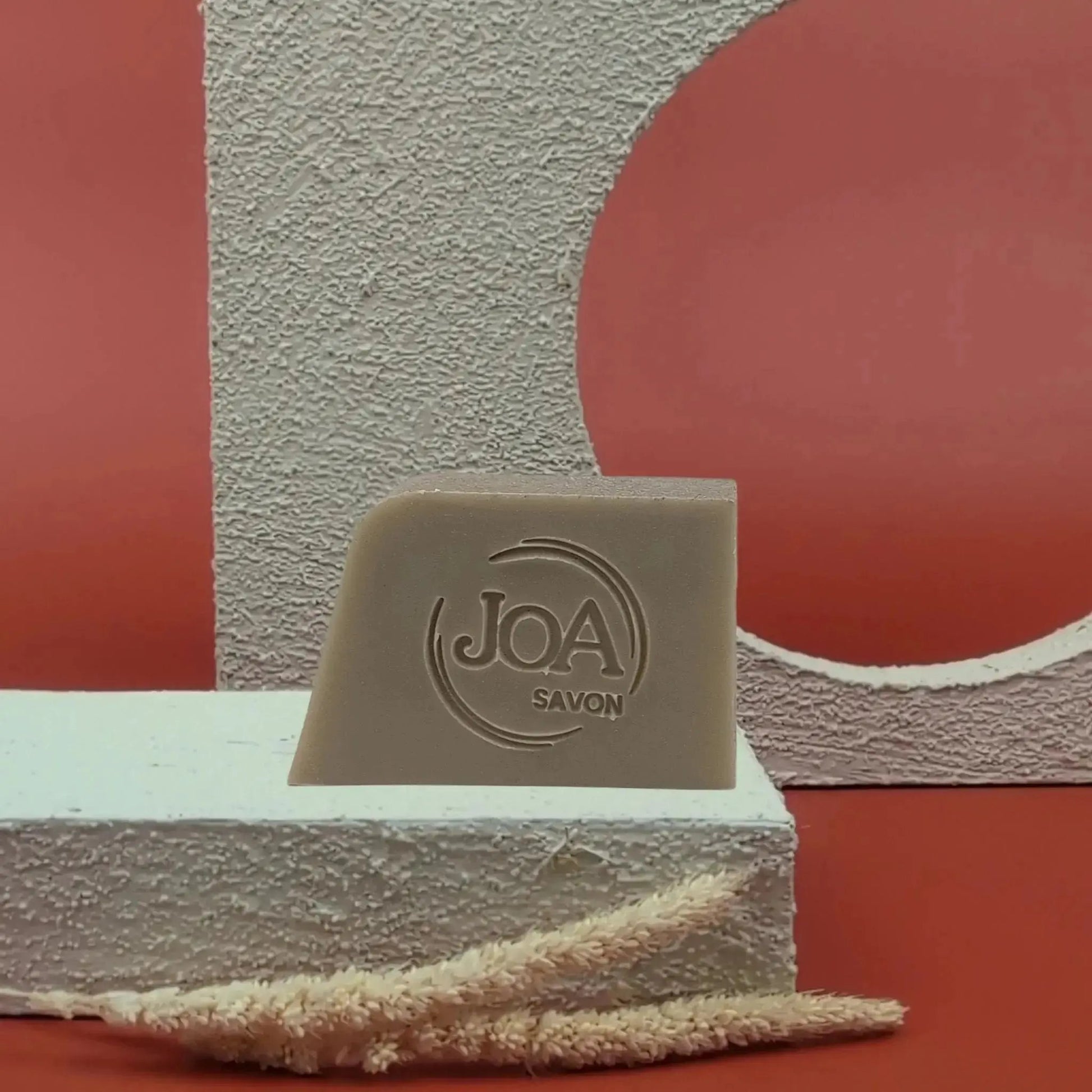 Savon solide Boue de la mer morte Joa beige-gris, posé sur socle blanc texturé avec fond rouge et structures géométriques