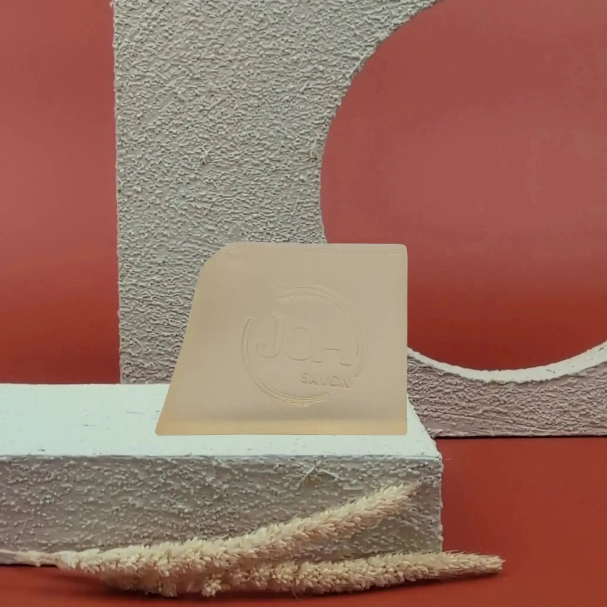 Savon solide Calendula Joa beige doux, posé sur socle blanc texturé avec fond rouge et éléments géométriques