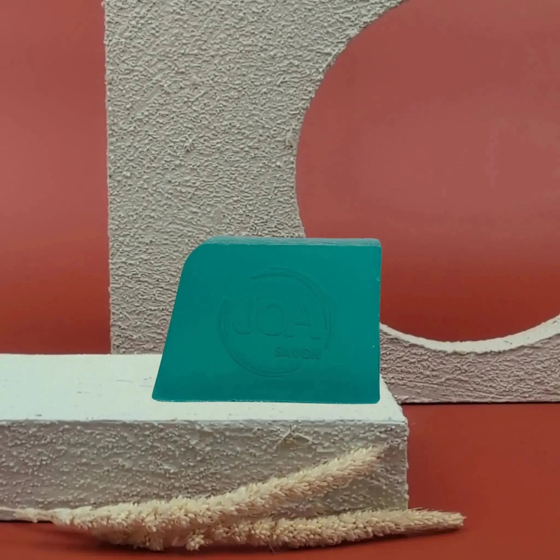 Savon solide Chanvre Joa vert profond, posé sur socle blanc texturé avec fond rouge et formes géométriques