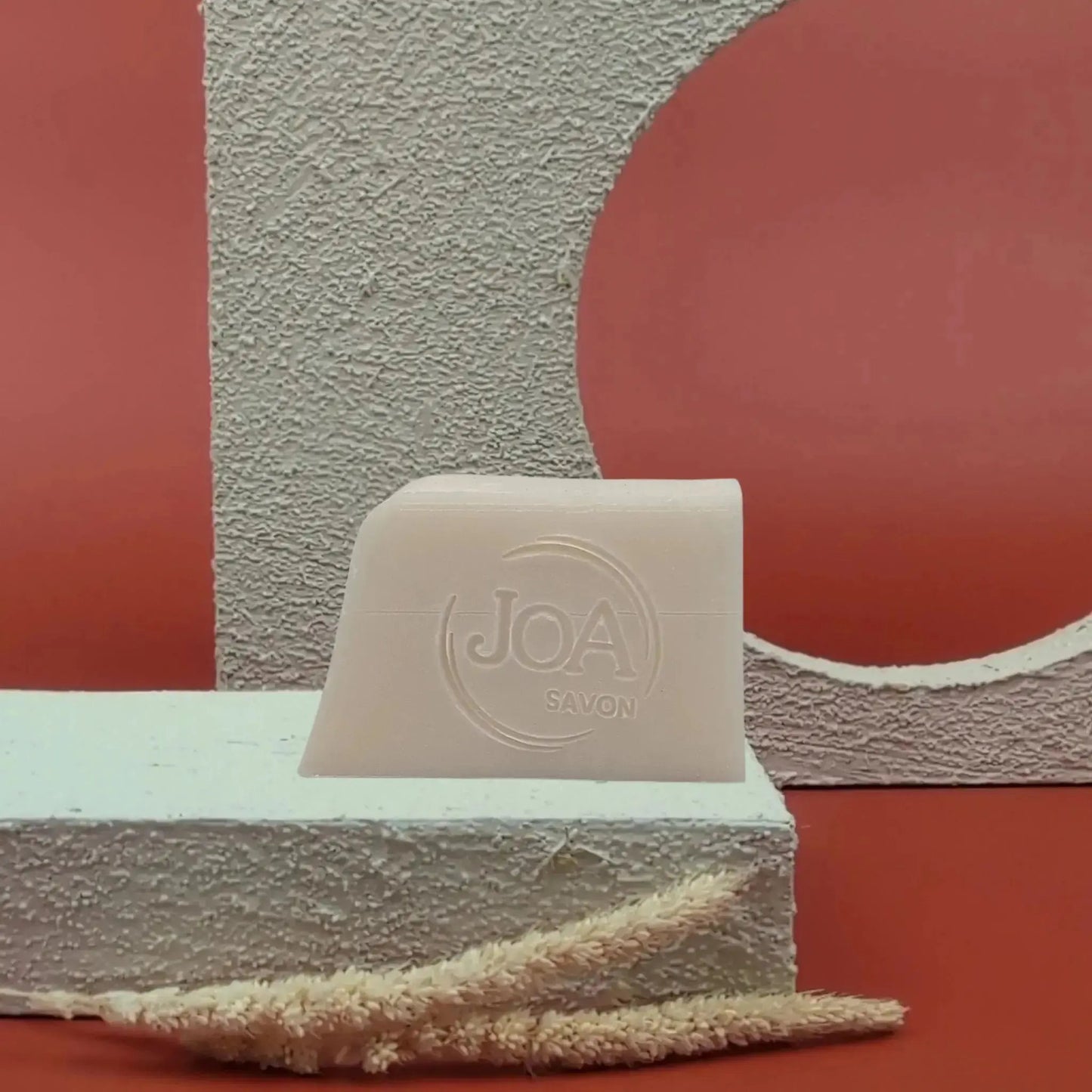 Savon solide Coco Joa beige clair, posé sur socle blanc texturé avec fond rouge et structure géométrique