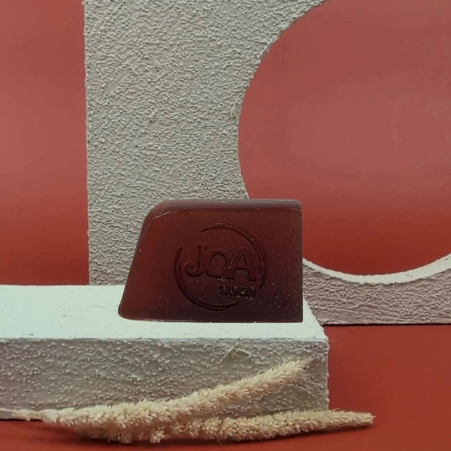 Savon solide Coco Vanille Joa brun intense, posé sur socle blanc texturé avec fond rouge et structure géométrique