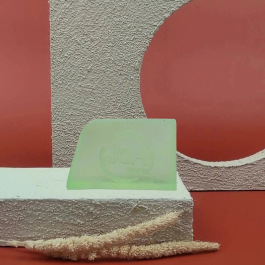 Savon solide Concombre Joa vert pastel, posé sur socle blanc texturé avec fond rouge et éléments géométriques