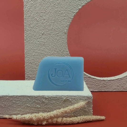 Savon solide Corail Sport Joa bleu givré, posé sur socle blanc texturé avec fond rouge et structure géométrique