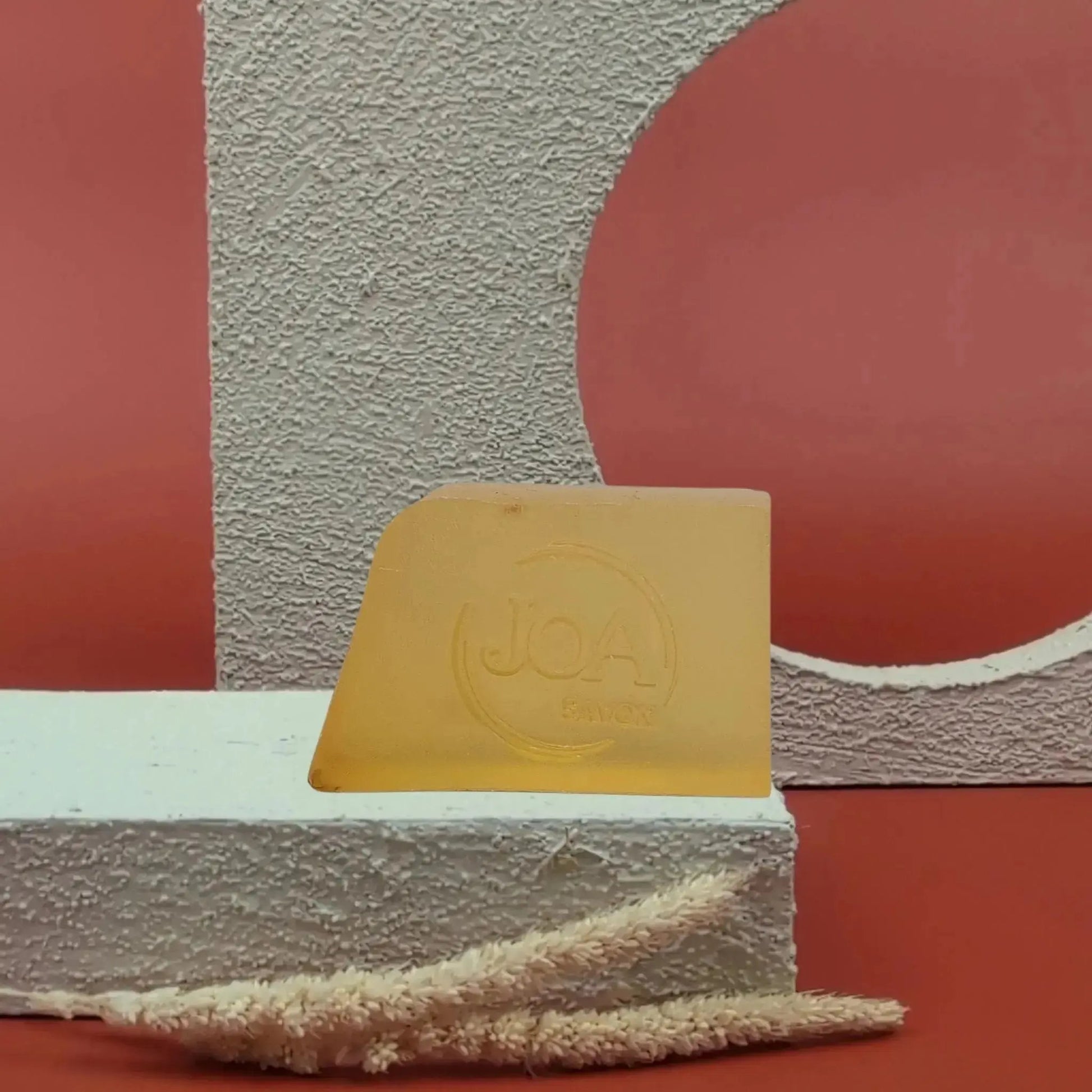 Savon solide Fleur d’Oranger Joa jaune doré, posé sur socle blanc texturé avec fond rouge et structure géométrique