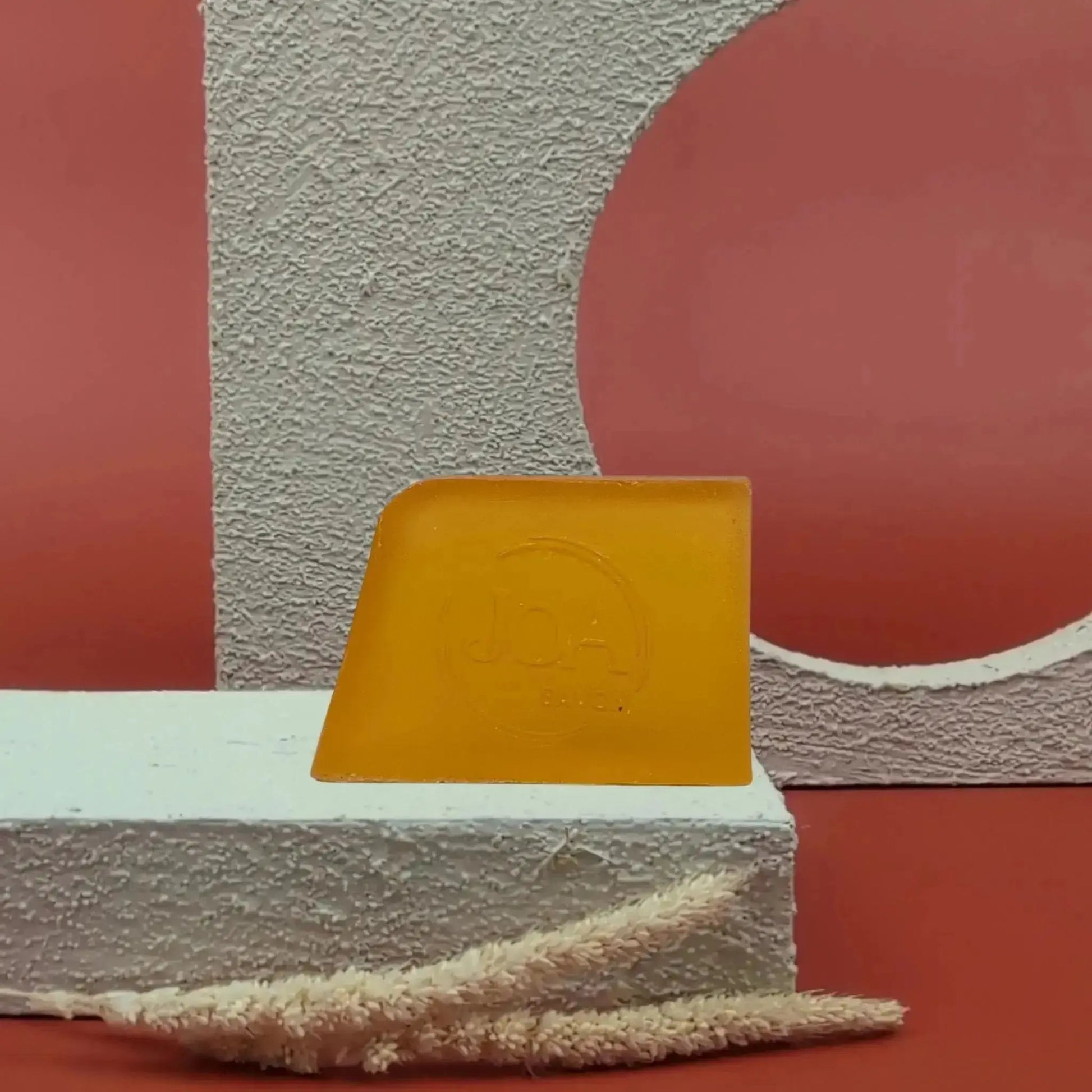 Savon solide Frangipanier Joa jaune orangé, posé sur socle blanc texturé avec fond rouge et structure géométrique