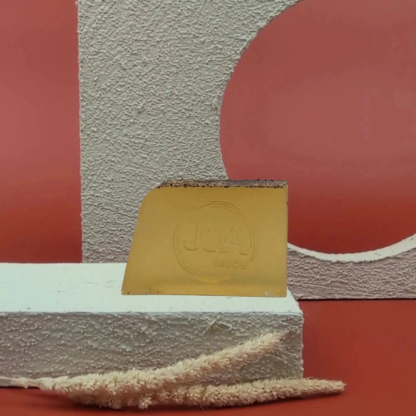 Savon solide Fruit de la Passion Joa jaune doré, posé sur socle blanc texturé avec fond rouge et structure géométrique