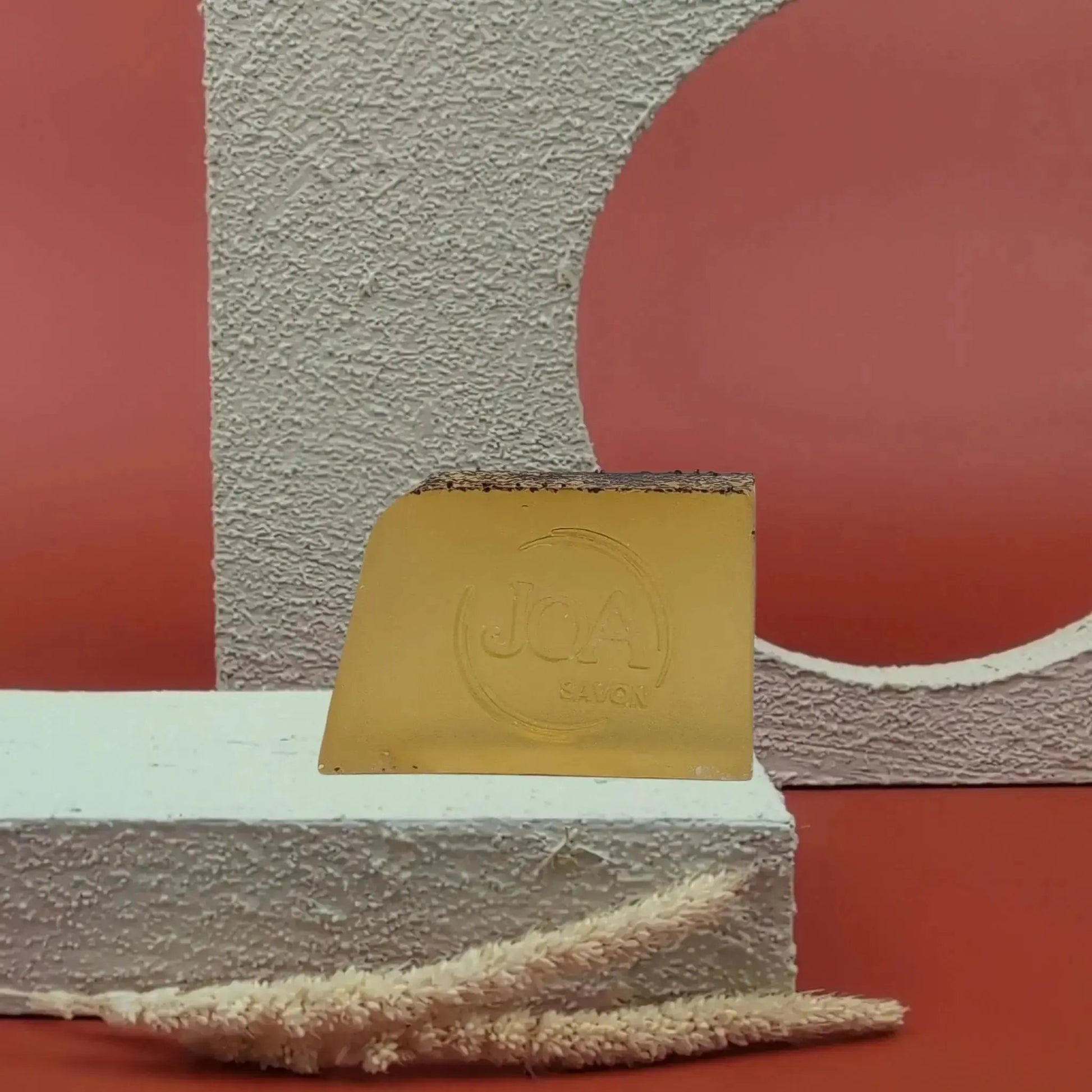 Savon solide Fruit de la Passion Joa jaune doré, posé sur socle blanc texturé avec fond rouge et structure géométrique