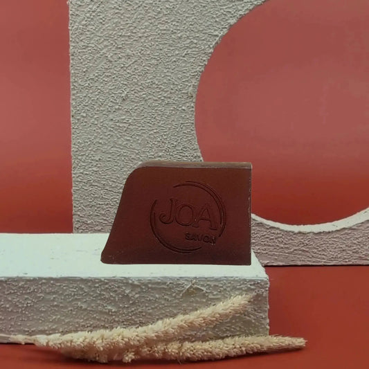 Savon solide Havane Vanille Joa brun profond, posé sur socle blanc texturé avec fond rouge et structure géométrique