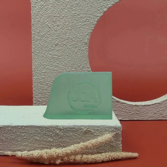 Savon solide L’homme Joa vert doux, posé sur socle blanc texturé avec fond rouge et structure géométrique