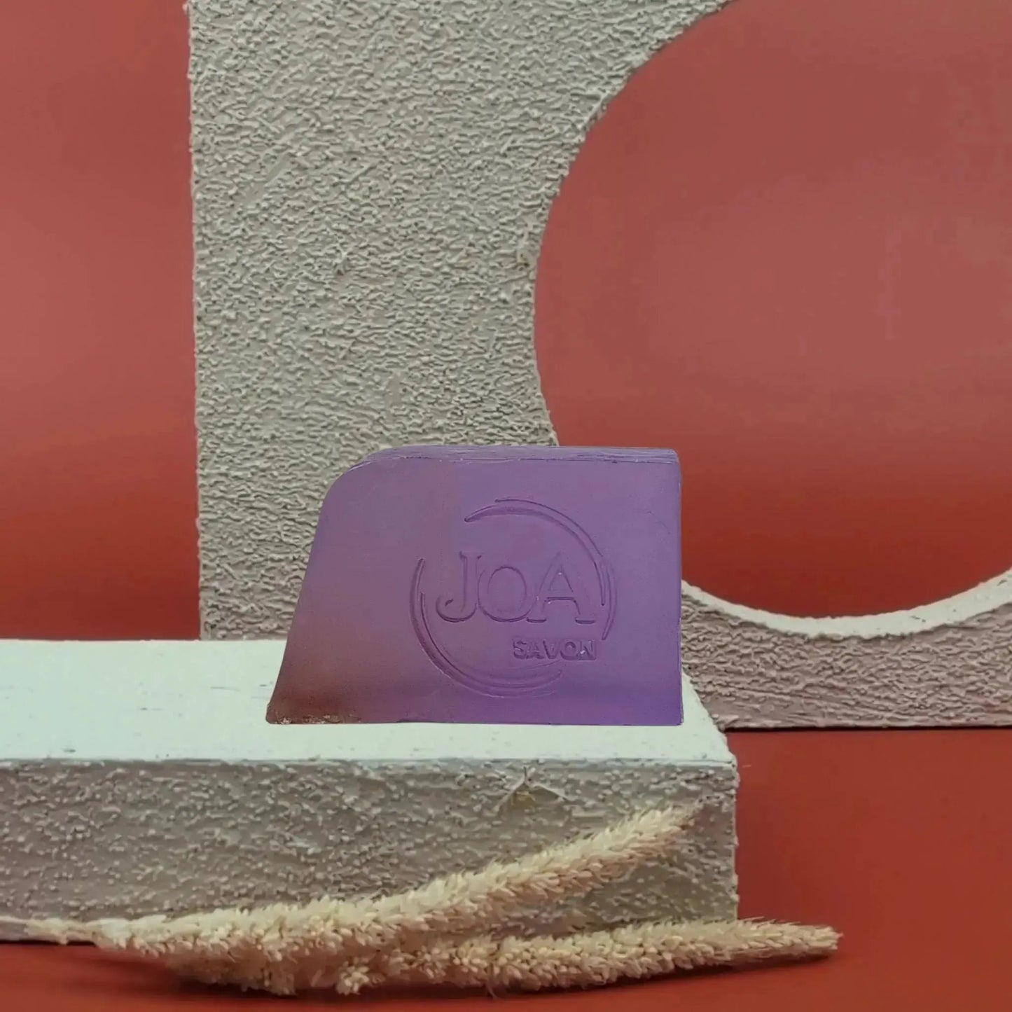 Savon solide Lavande Joa de couleur violette, posé sur socle blanc texturé avec fond rouge et structure géométrique