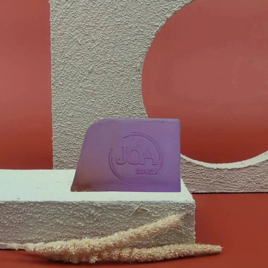 Savon solide Lavande Joa de couleur violette, posé sur socle blanc texturé avec fond rouge et structure géométrique
