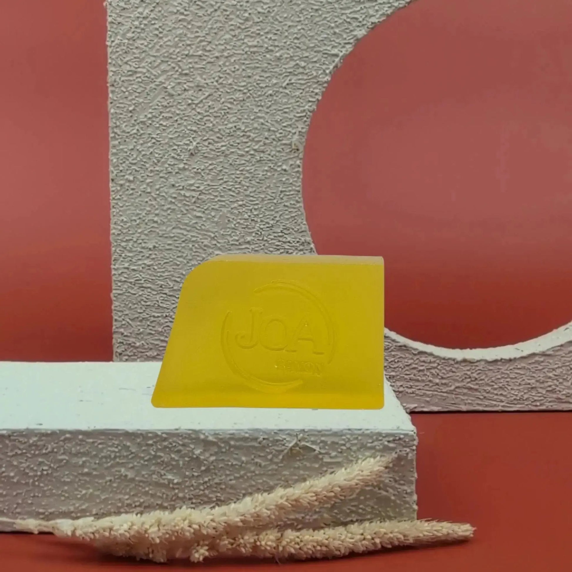 Savon solide Mangue Joa jaune lumineux, posé sur socle blanc texturé avec fond rouge et structure géométrique