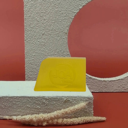 Savon solide Mangue Joa jaune lumineux, posé sur socle blanc texturé avec fond rouge et structure géométrique