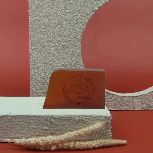 Savon solide Monoï Joa couleur ambrée, posé sur socle blanc avec fond rouge et cercle texturé