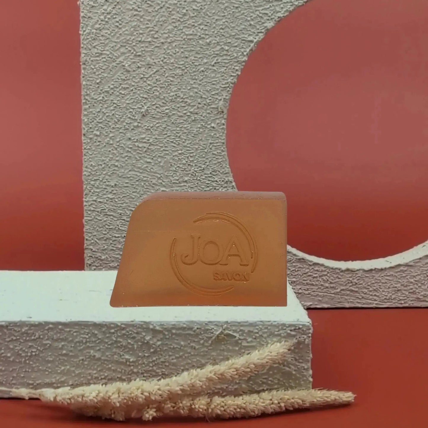 Savon solide Muxu Joa couleur caramel, présenté sur socle blanc avec fond rouge graphique