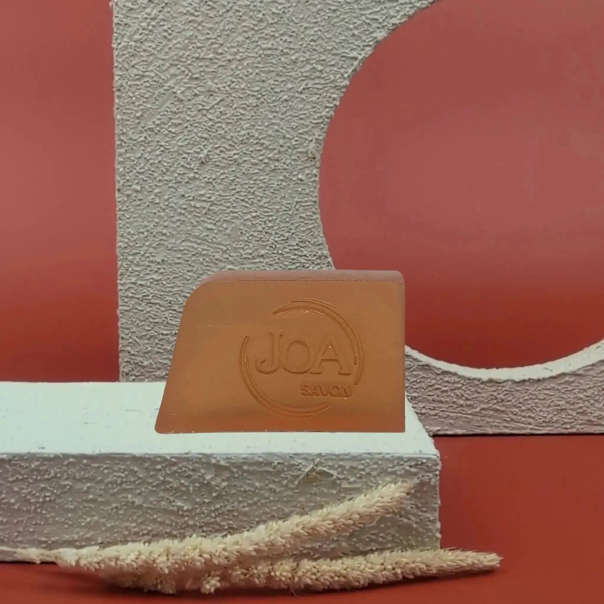 Savon solide Muxu Joa couleur caramel, présenté sur socle blanc avec fond rouge graphique