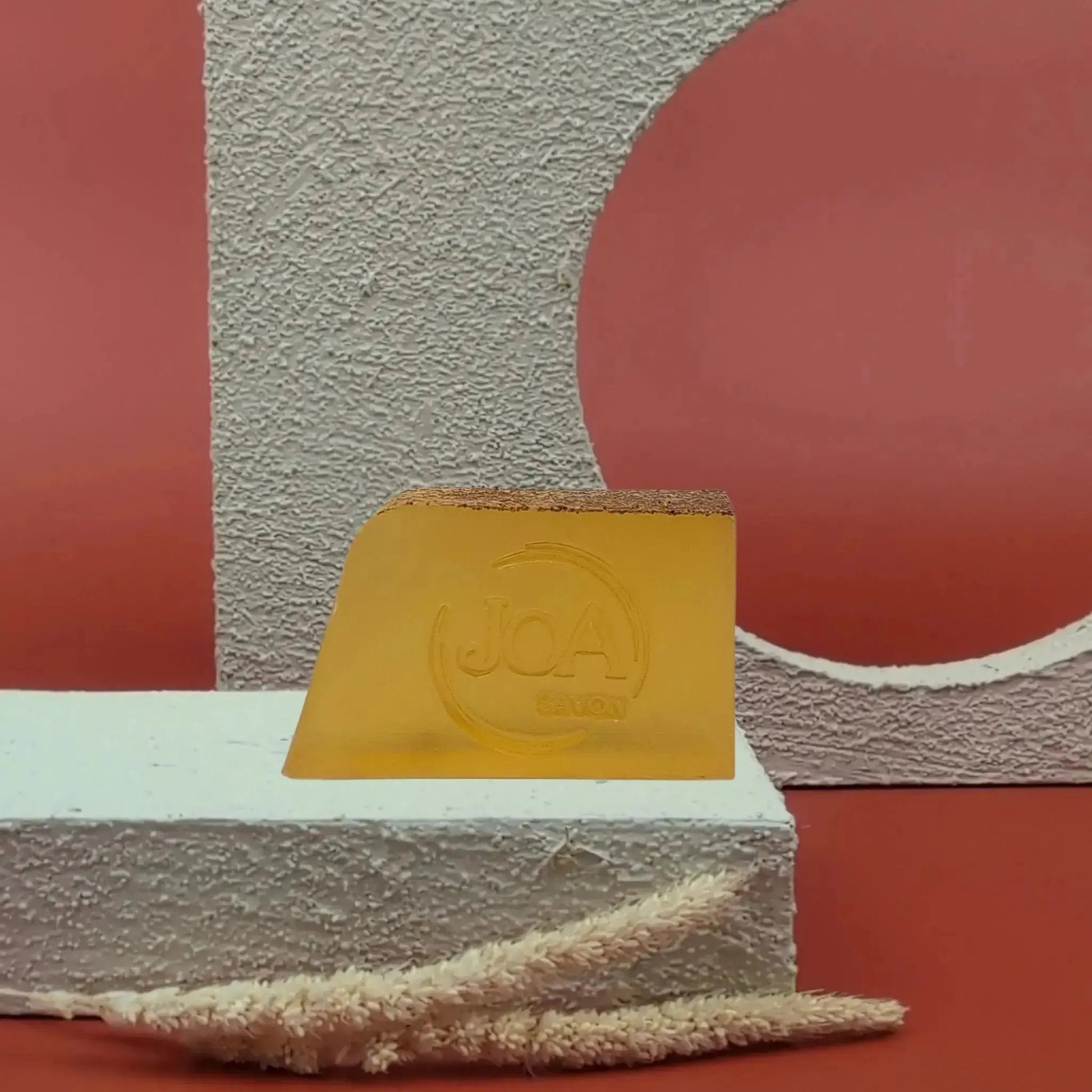 Savon solide Orange Pamplemousse Joa, orange vif translucide, sur fond rouge texturé et socle blanc moderne