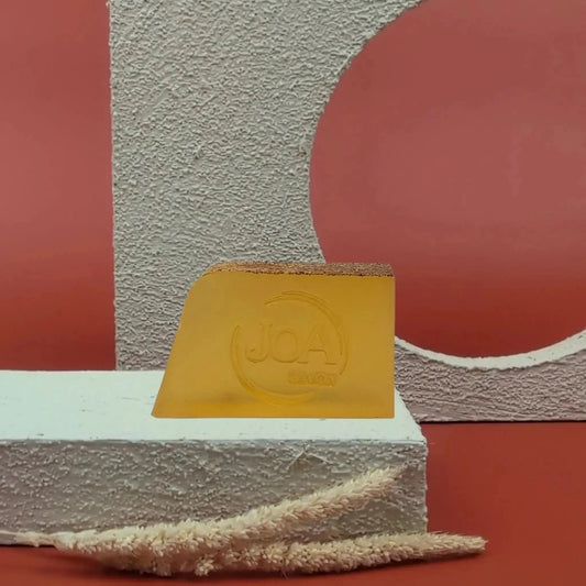 Savon solide Orange Pamplemousse Joa, orange vif translucide, sur fond rouge texturé et socle blanc moderne