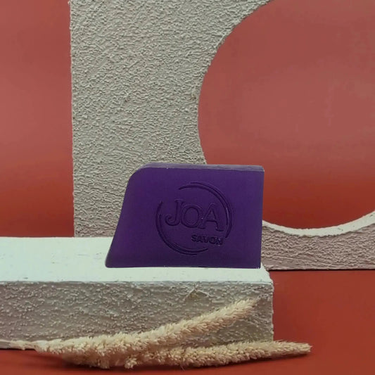 Savon solide Patchouli Joa, couleur violette intense, posé sur socle blanc, fond rouge texturé.