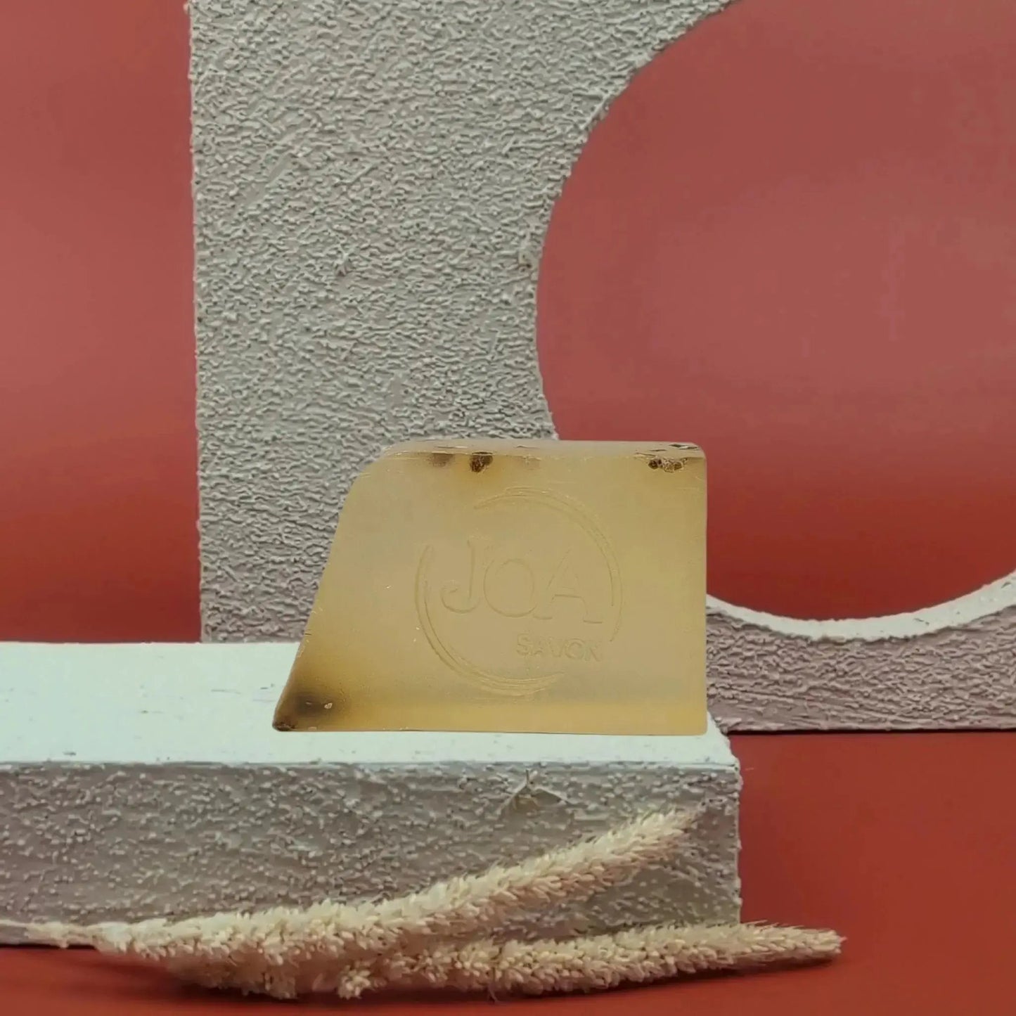 Savon solide Thé Vert de Joa, jaune transparent avec inclusions, posé sur socle blanc avec fond rouge texturé