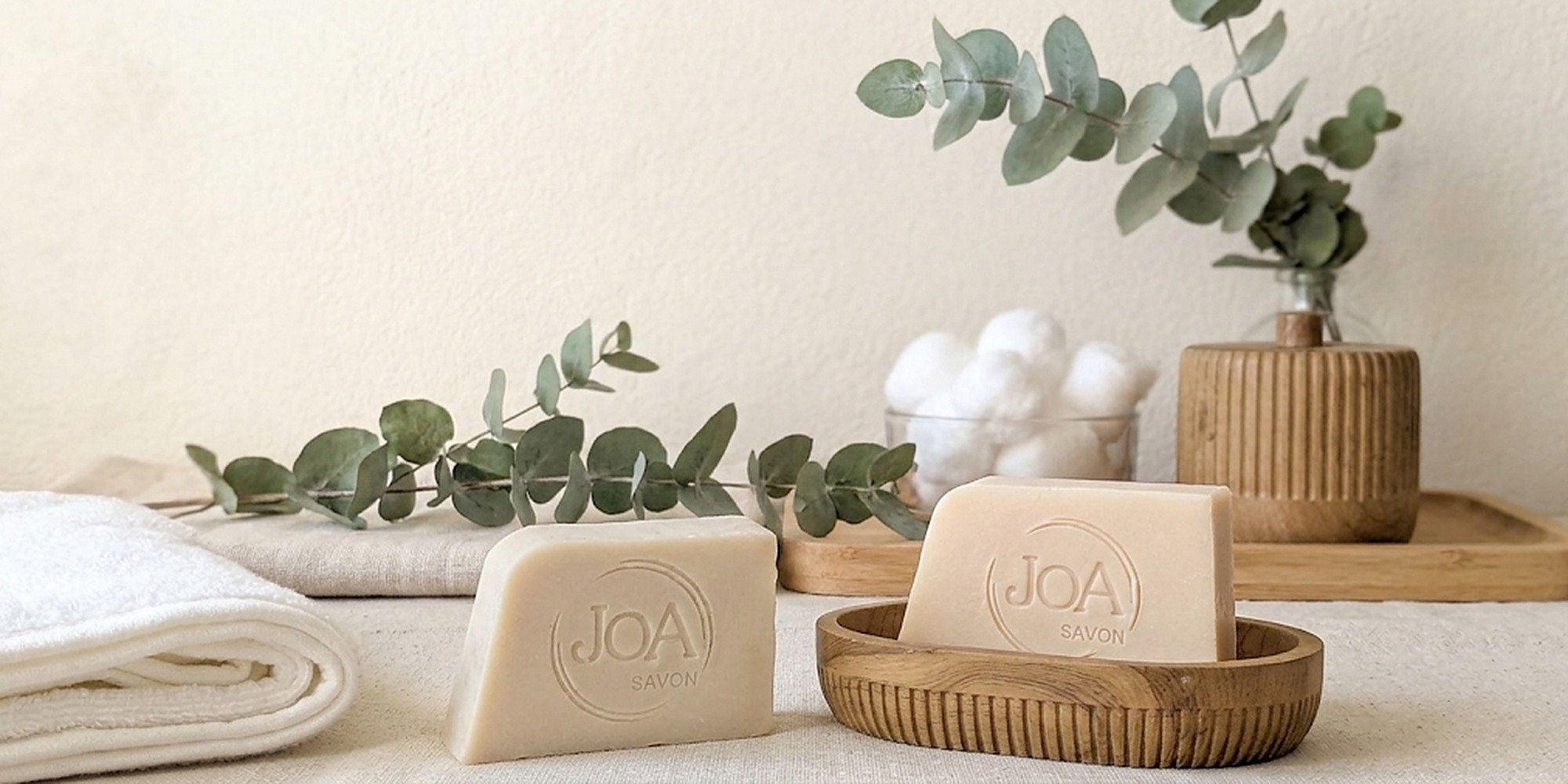 Deux savons Joa Savon crème et beige dans une salle de bain douce, avec une serviette, du coton et de l'eucalyptus