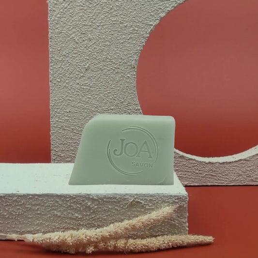 Savon solide Tea Tree de Joa, vert clair, posé sur un socle blanc avec décor texturé et fond rouge

