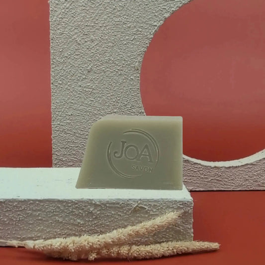 Savon solide Argile verte Joa vert grisé, posé sur socle blanc texturé avec fond rouge et structure géométrique

