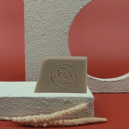 Savon solide Boue de la mer morte Joa beige-gris, posé sur socle blanc texturé avec fond rouge et structures géométriques

