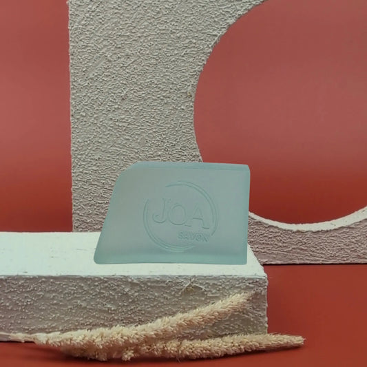 Savon solide Brise de mer Joa bleu clair, posé sur socle blanc texturé avec fond rouge et arc géométrique

