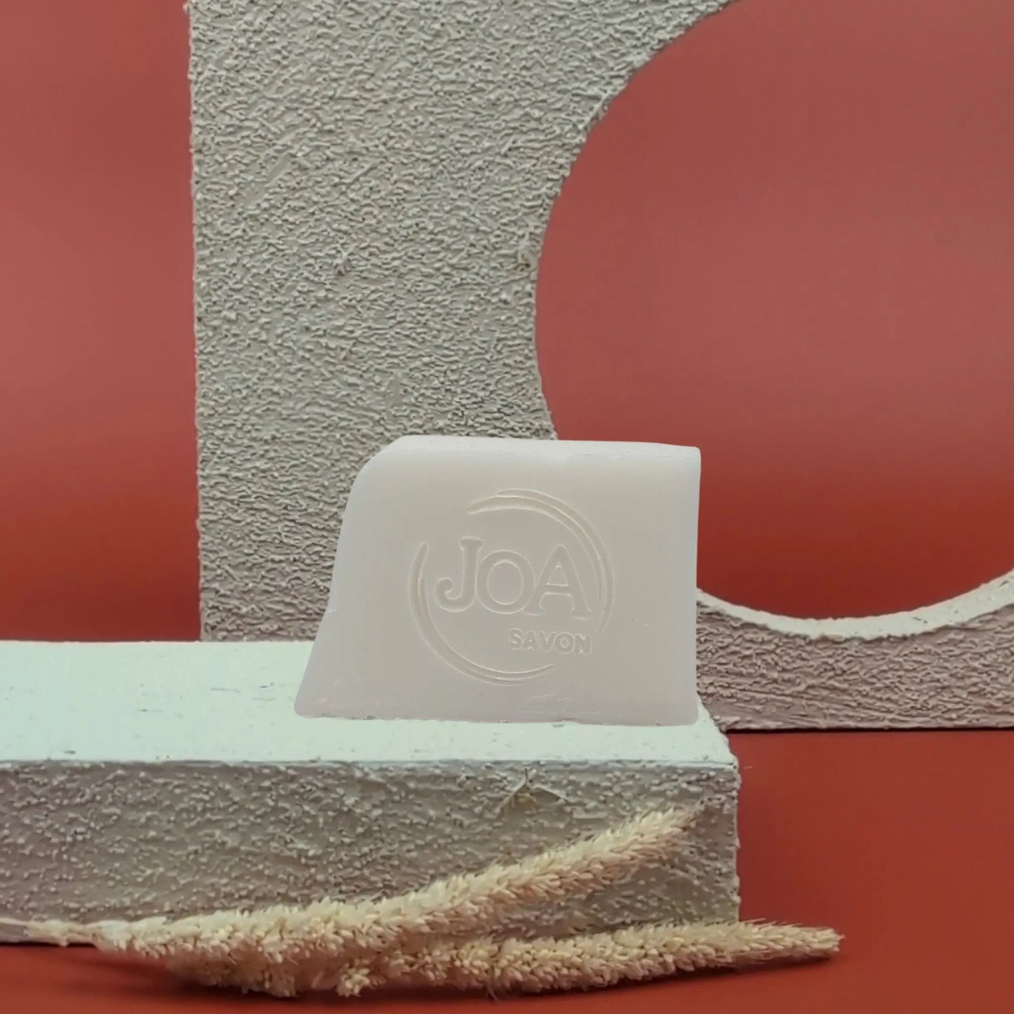 Savon solide Lait d’ânesse Joa ivoire clair, posé sur socle blanc texturé avec fond rouge et structure géométrique

