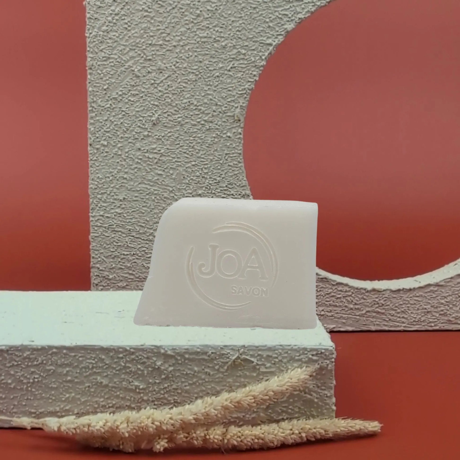 Savon solide Lait d’ânesse Joa ivoire clair, posé sur socle blanc texturé avec fond rouge et structure géométrique

