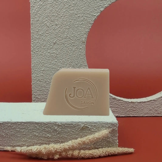 Savon solide Lait de figue Joa couleur beige, posé sur socle blanc texturé avec fond rouge et formes géométriques

