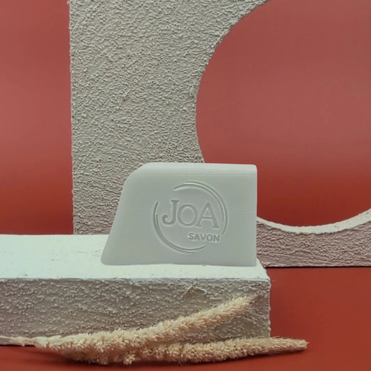 Savon solide Marine Joa bleu très pâle, posé sur socle blanc texturé avec fond rouge et décor géométrique en arrière-plan

