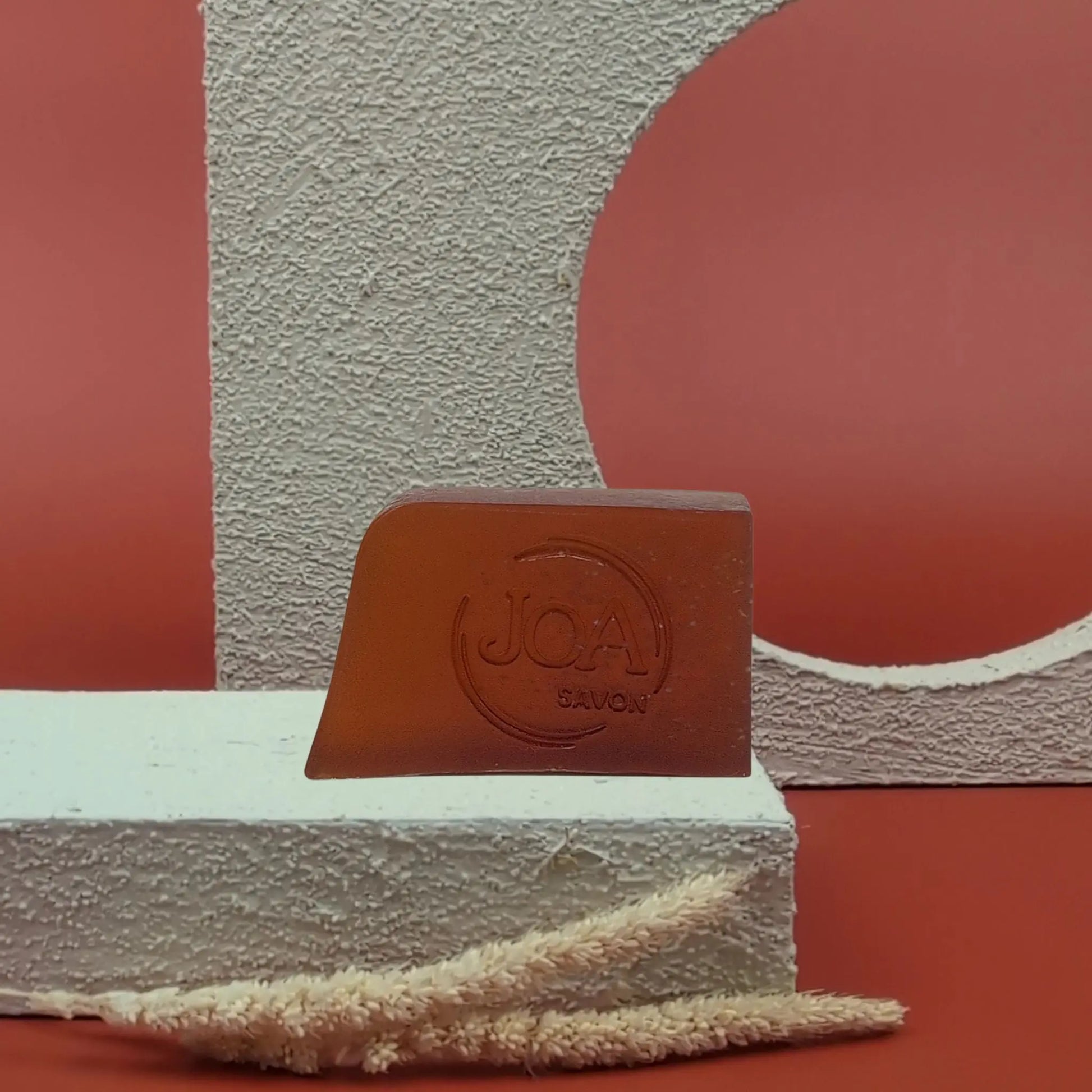 Savon solide Monoï Joa couleur ambrée, posé sur socle blanc avec fond rouge et cercle texturé

