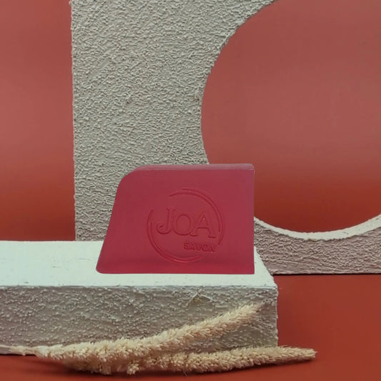 Savon solide Oud Joa, couleur rouge profond, posé sur socle texturé blanc avec fond rouge moderne

