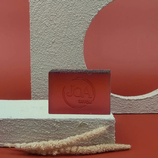 Savon solide Porto Joa, couleur rouge foncé, posé sur un socle blanc, avec fond rouge texturé.
