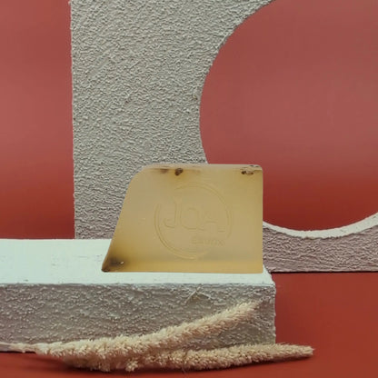 Savon solide Thé Vert de Joa, jaune transparent avec inclusions, posé sur socle blanc avec fond rouge texturé

