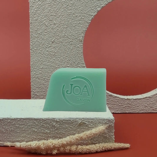 Savon solide Verveine Basilic de Joa, vert pastel, posé sur socle blanc avec fond rouge texturé

